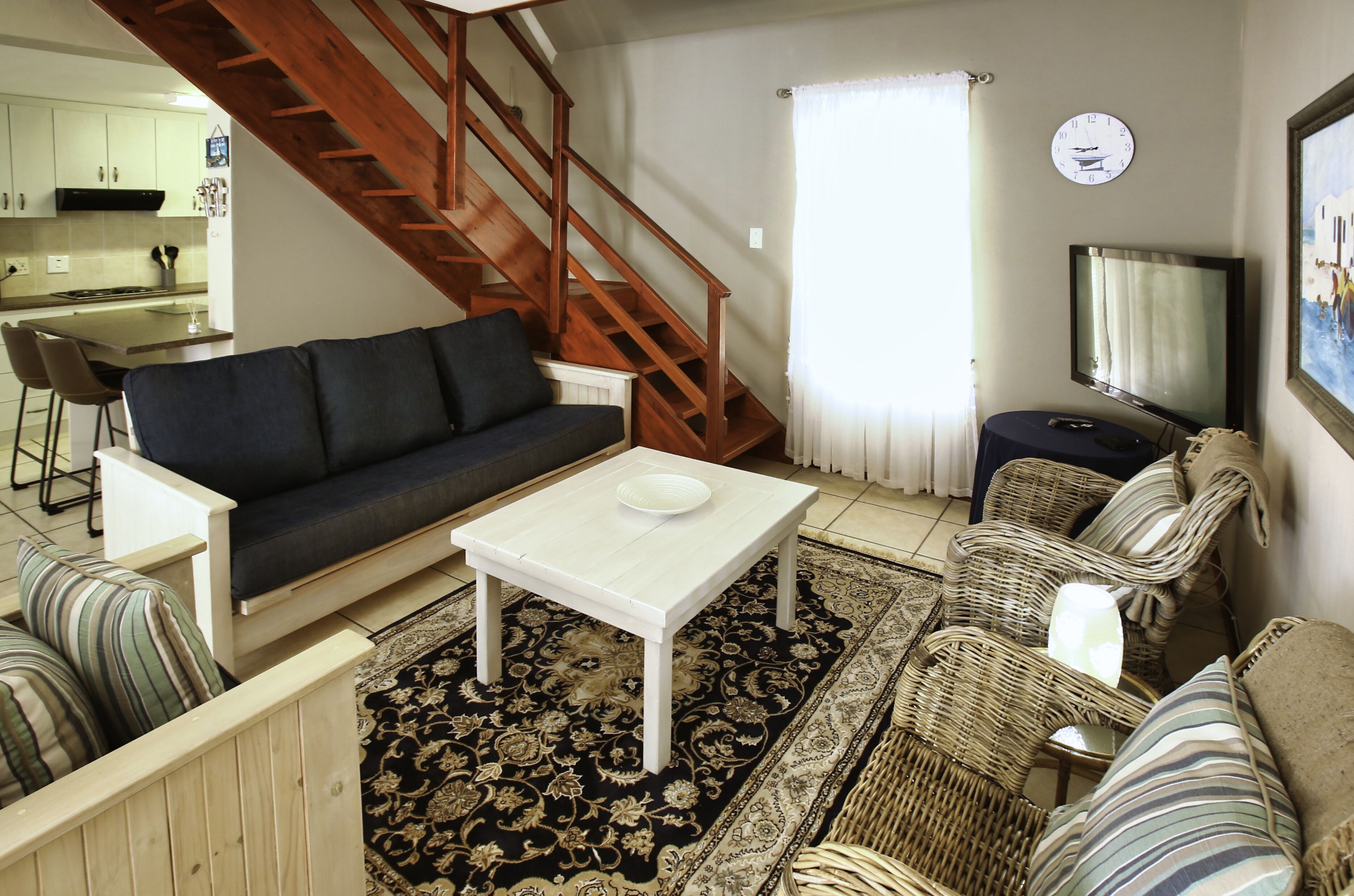 A living room with a sofa, coffee table, and wicker chairs, a staircase, a kitchen in the background, and a clock on the wall.