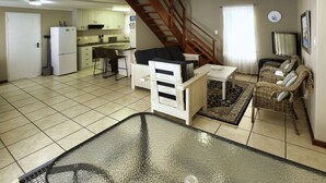A spacious living area with a kitchen, a dining table, a sofa, a glass-top coffee table, and a staircase leading to an upper level.