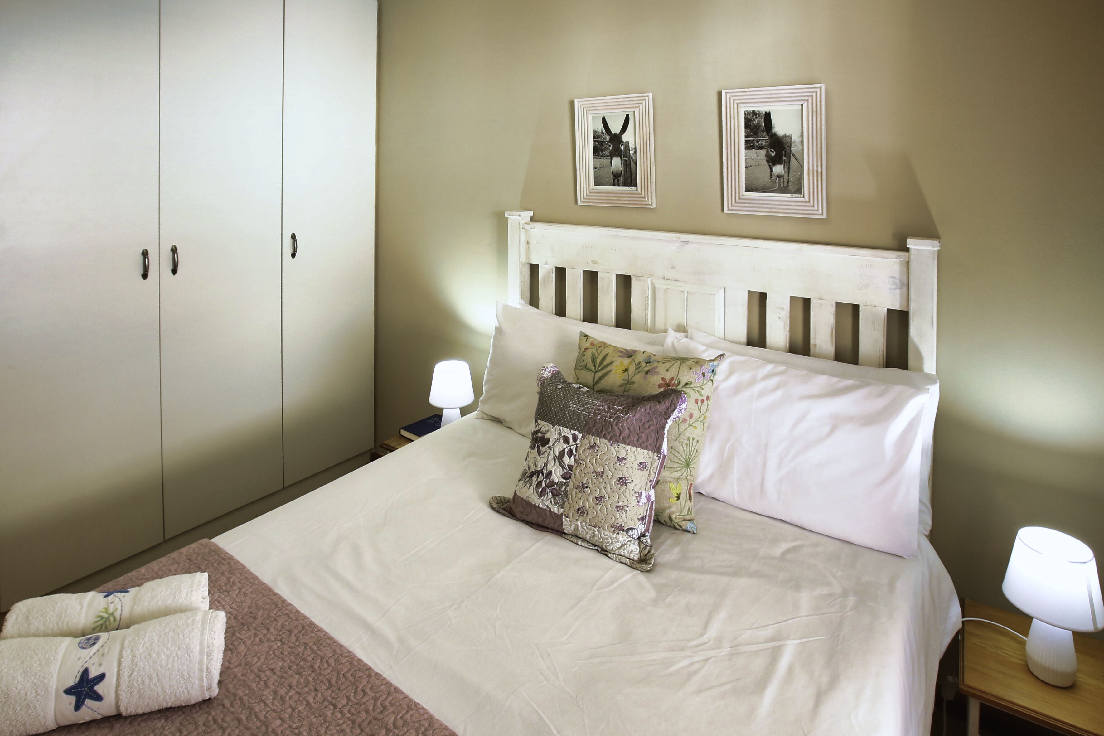 A neatly made bed with white linens and decorative pillows, a wooden nightstand with a lamp, and a wardrobe with a handle.