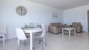 Un salon moderne avec une table à manger, des chaises blanches, un canapé beige et une horloge murale ronde.