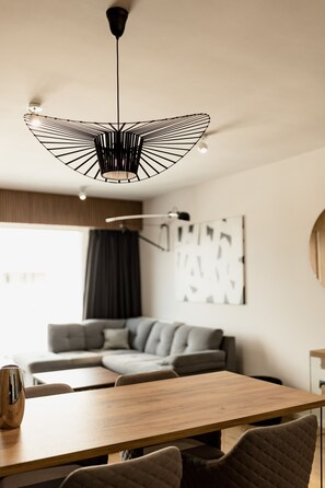 A modern dining area with a wooden table and chairs, a unique ceiling light fixture, and a gray sofa in the background. - Nest Inn Mirror Apartment (Cluj-Napoca)