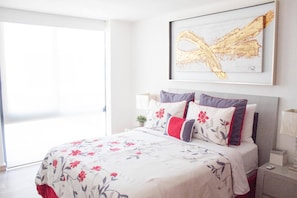 Un dormitorio con cama, almohadas, mesita de noche, lámpara y una obra de arte enmarcada en la pared.