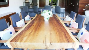 Una larga mesa de comedor de madera con sillas, un florero con flores y una cocina al fondo.