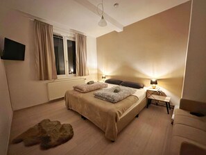 Un dormitorio con una cama grande, un televisor de pantalla plana en la pared, una ventana con cortinas, un radiador y una pequeña mesa con una lámpara.