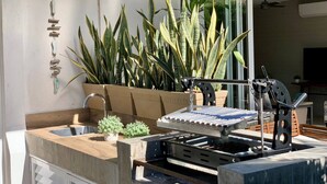 Une cuisine extérieure moderne, dotée d’un barbecue encastré, d’un évier et d’un bac à plantes où sont disposées de grandes plantes vertes.