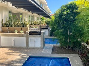 Un espace piscine agrémenté d’un barbecue intégré, de plantes en pot et d’un coin salon extérieur abrité.