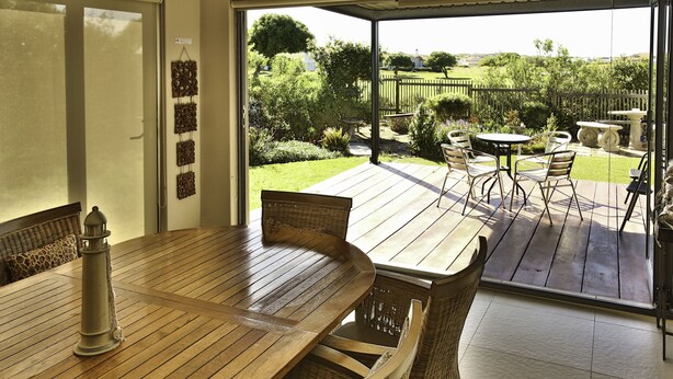 A wooden deck with a table and chairs, a wicker chair, and a view of a garden with a fence and trees.