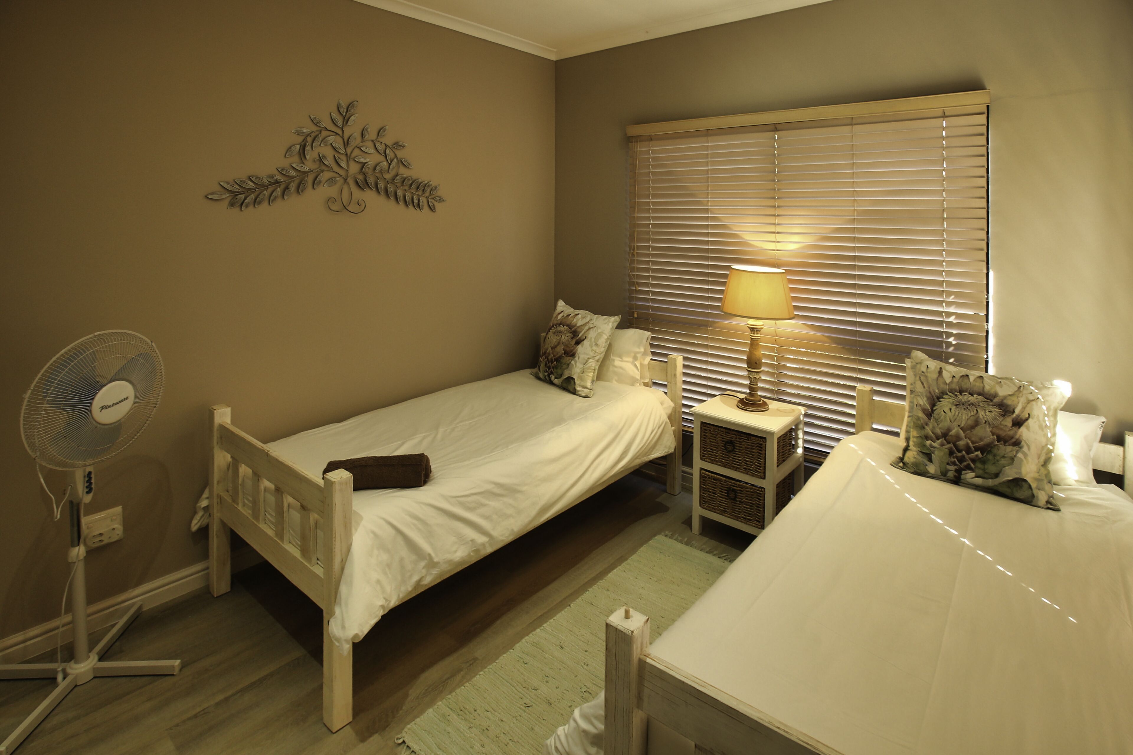 Een slaapkamer met een enkel bed, een nachtkastje met een lamp, een ventilator aan de muur en een raam met zonwering.