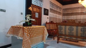 A room with a wooden door, a table with a patterned tablecloth, a sofa with a patterned cushion, and lace curtains. - OYO 93706 Penginapan Syariah Hj. Kenan (Koto Tinggi)