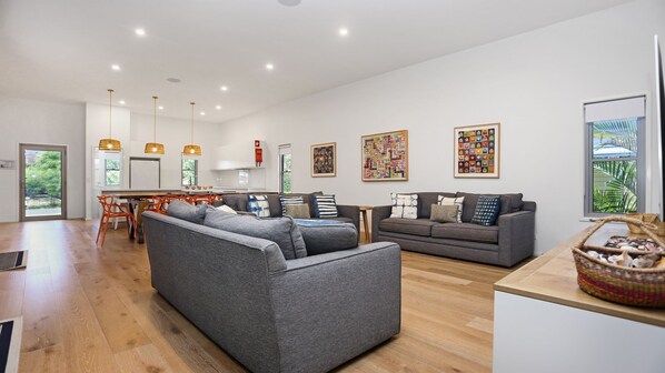 A spacious living area with a grey sofa, a wooden dining table, and a kitchen in the background. - Russell Street Retreat (Hawks Nest)
