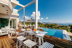 Un espace piscine extérieur moderne, doté d’une terrasse en bois, de mobilier blanc et offrant une vue panoramique sur les montagnes et la mer.
