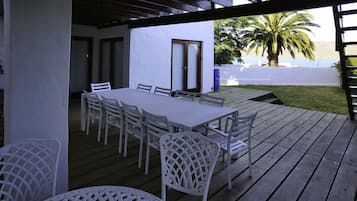 Un patio techado al aire libre con una mesa y sillas blancas, una terraza de madera y vistas al mar y palmeras.