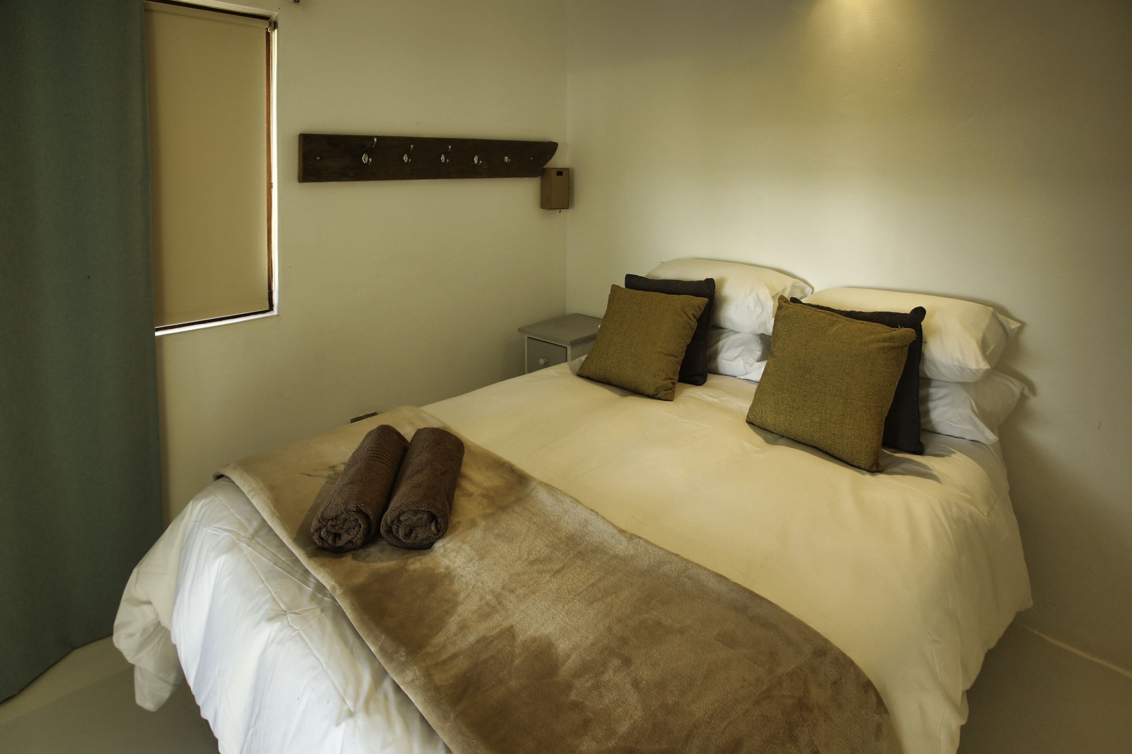 A neatly made bed with a beige comforter and two rolled towels, a small bedside table with a lamp, and a wall-mounted shelf above the bed.