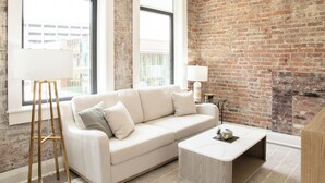 Un salon moderne avec un canapé blanc, une table basse et une lampe. On y trouve un mur en briques et une fenêtre donnant sur des immeubles.