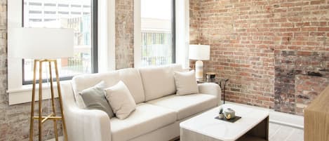 A modern living room with a white sofa, a coffee table, and a lamp. There is a brick wall and a window with a view of buildings.