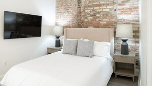 Une chambre avec un mur en briques, un lit avec une literie blanche, deux tables de chevet avec des lampes et un téléviseur à écran plat fixé au mur.