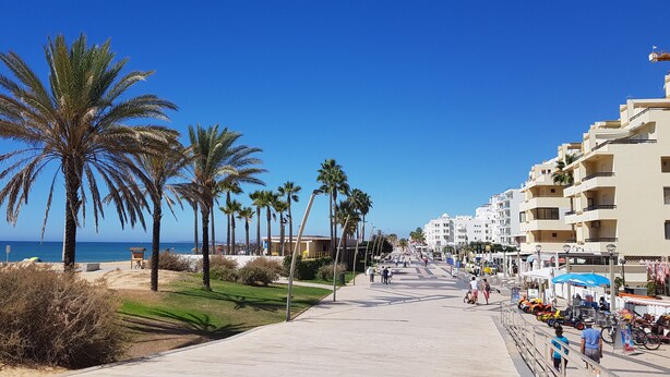 A coastal promenade with palm trees, a beach, and seaside buildings. - Apartamento T1 em Quarteira! (Quarteira)