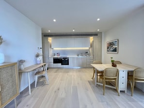 Een moderne keuken met ingebouwde apparatuur, een eethoek met een houten tafel en stoelen, en een klein zithoekje met een bank.