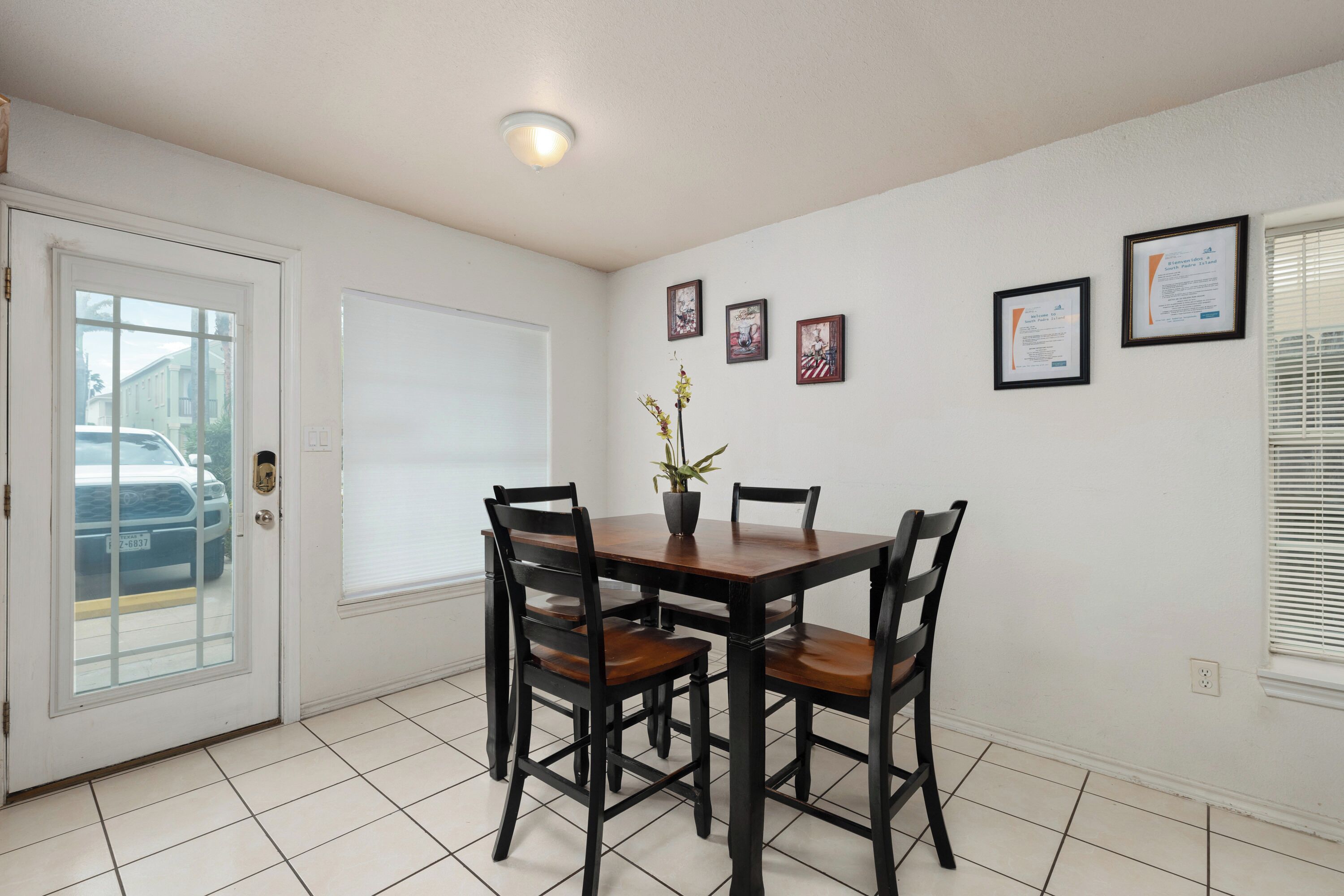 A dining area with a wooden table and four chairs, a potted plant, and framed certificates on the wall.