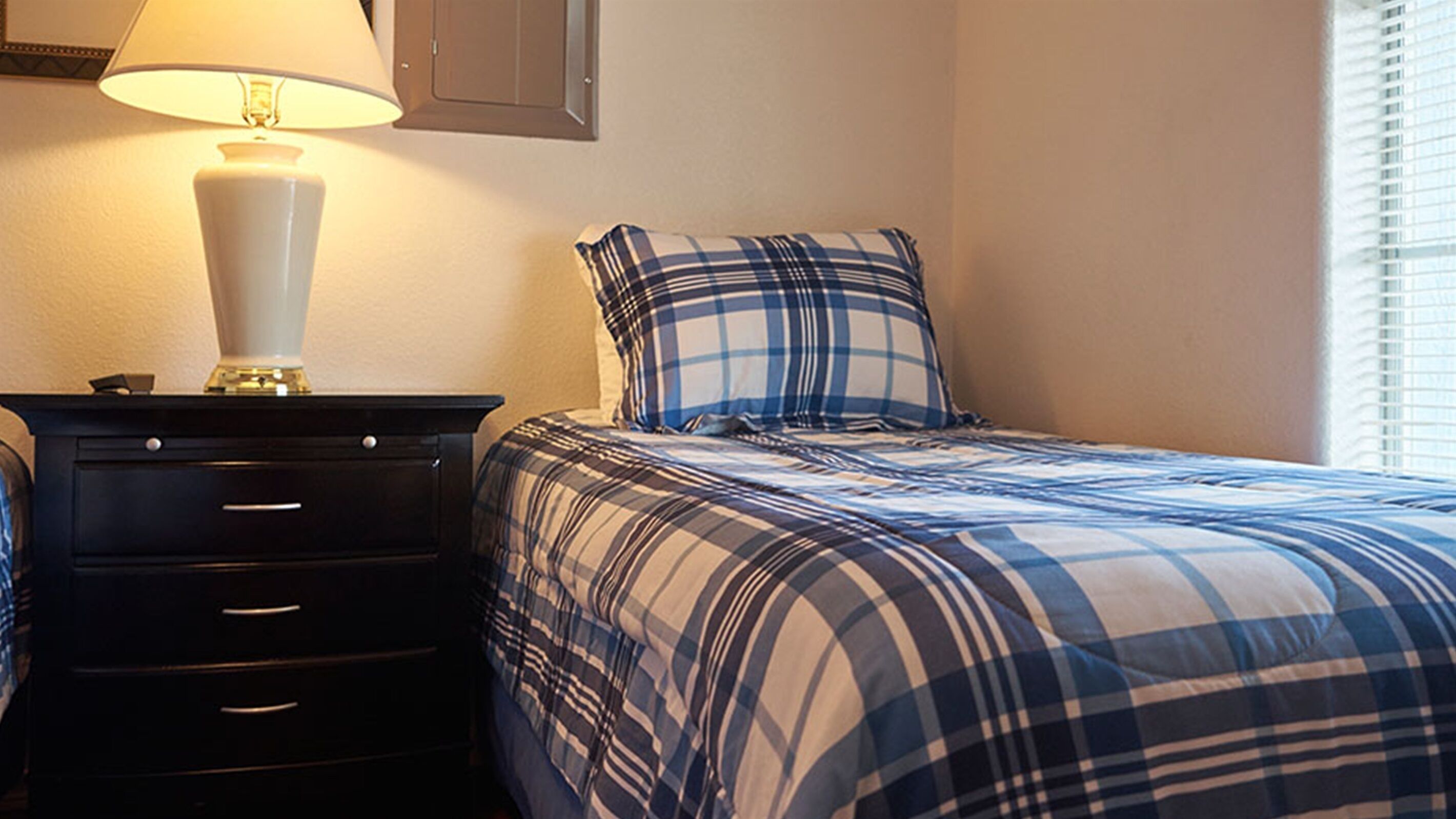 A bedroom with a bed, a nightstand, a lamp, and a window with blinds.