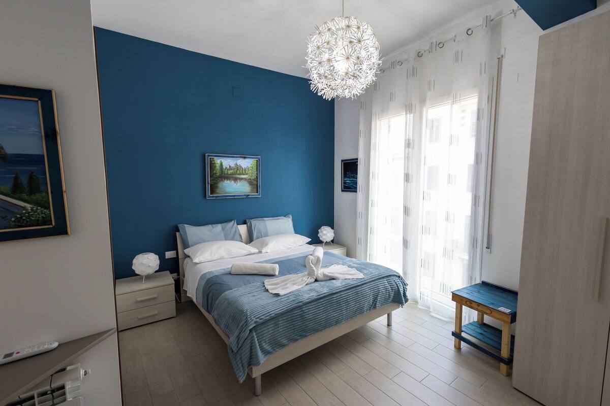 A bedroom with a bed, bedside tables, a nightstand, a wooden bench, and a large window with curtains.