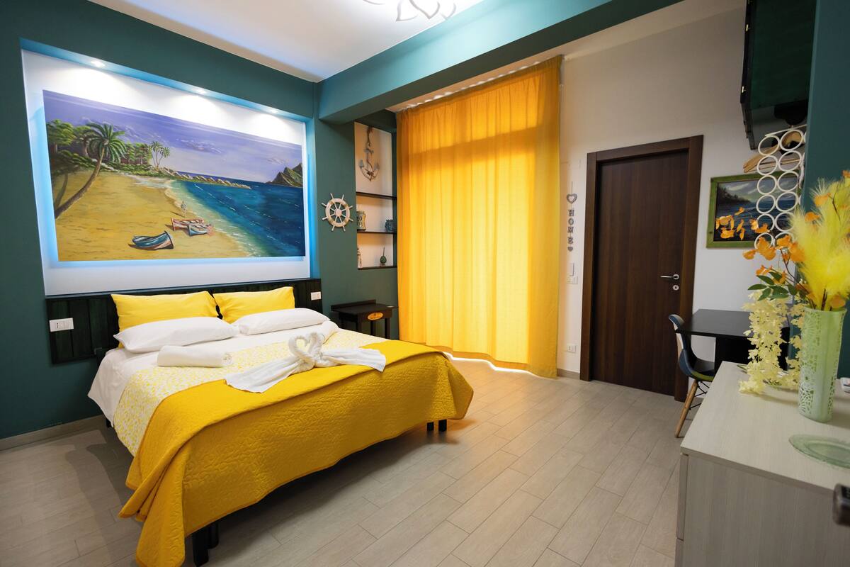 A bedroom with a bed, a large painting of a beach, a window with curtains, and a desk with a vase of flowers.