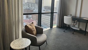 A modern hotel room with a large window, a comfortable armchair, a small round table, and a desk with a chair. - Neighbourgood Loop (Cape Town)