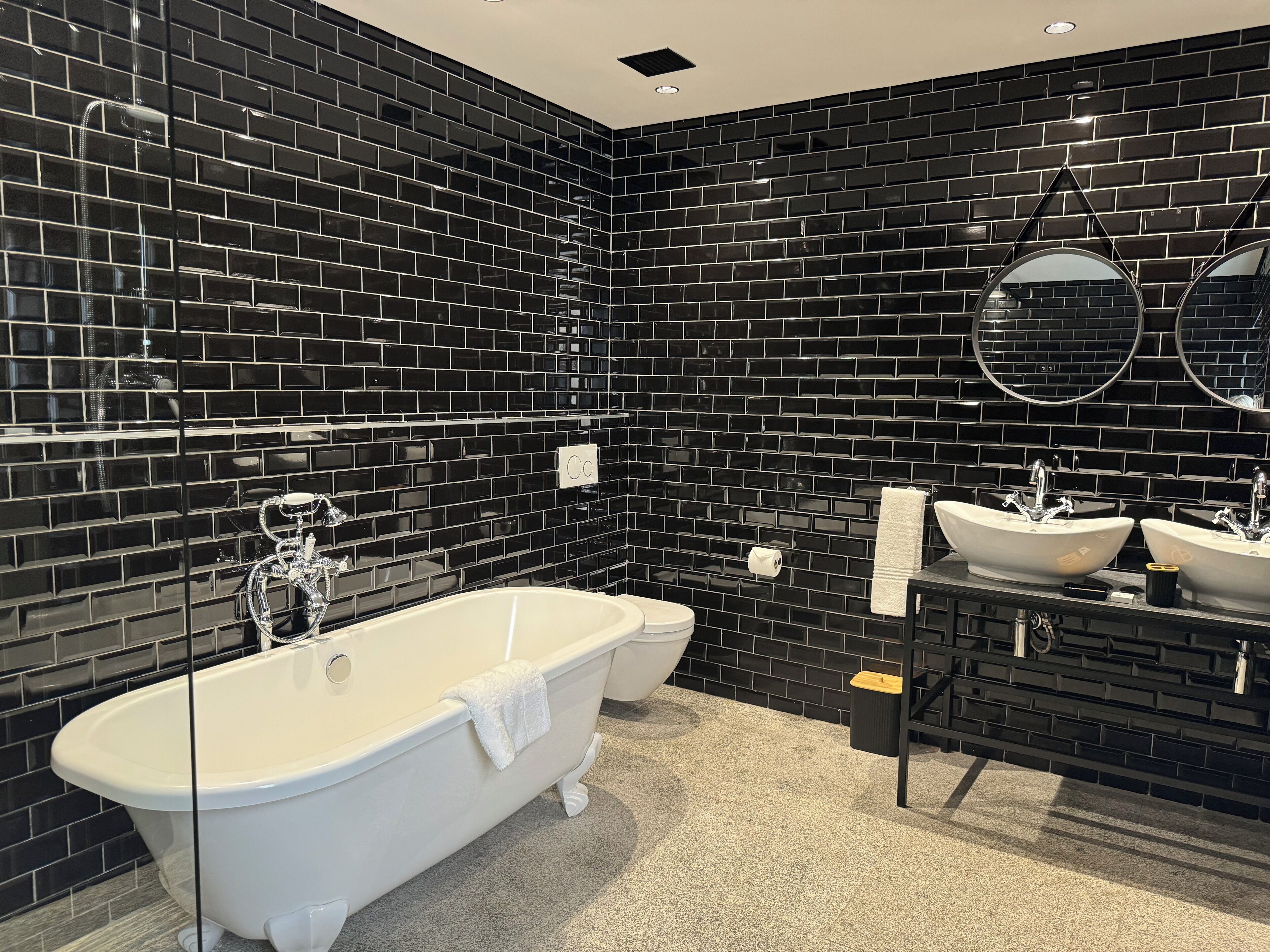 A modern bathroom with black tiled walls, a freestanding bathtub, two sinks, and a toilet.
