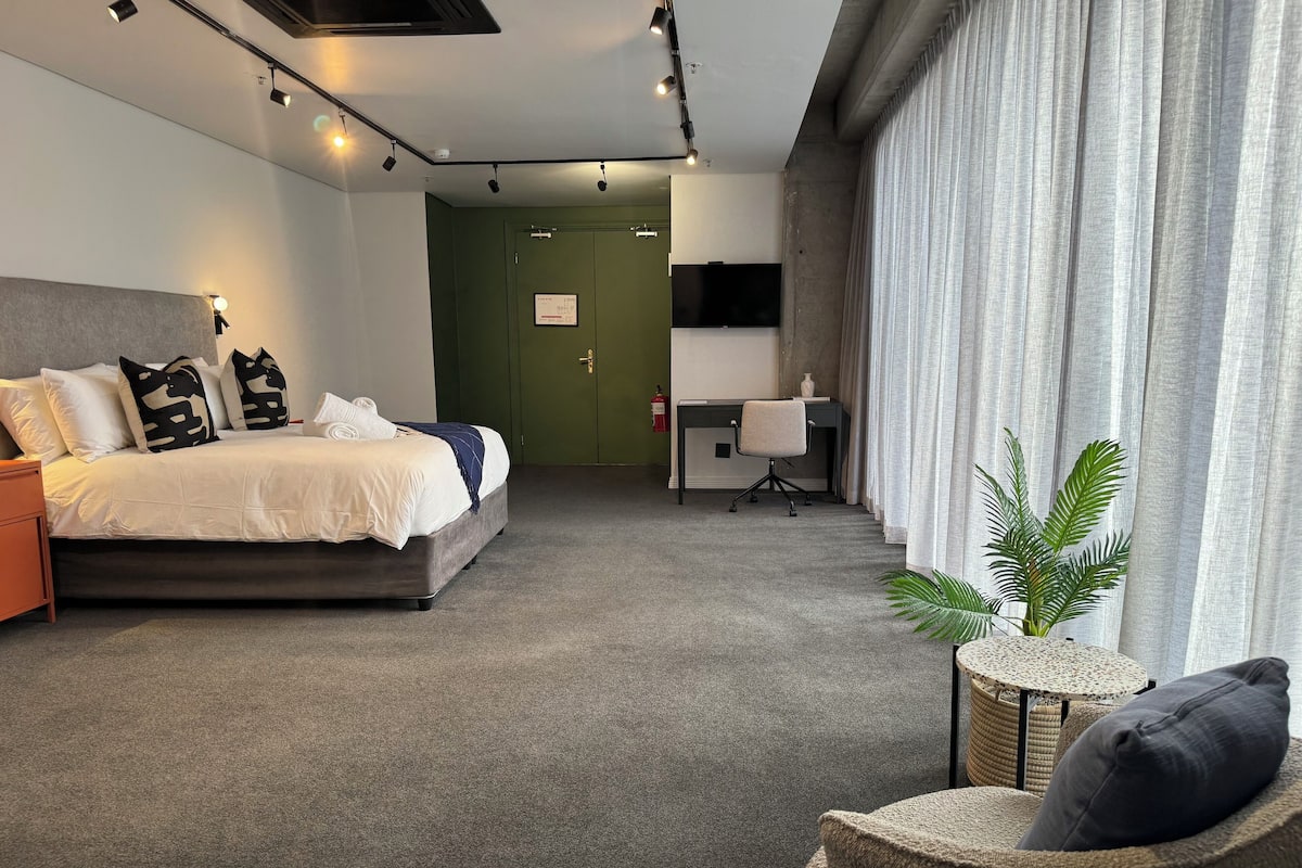 Quarto de hotel moderno com cama, escrivaninha, cadeira e planta em vaso.