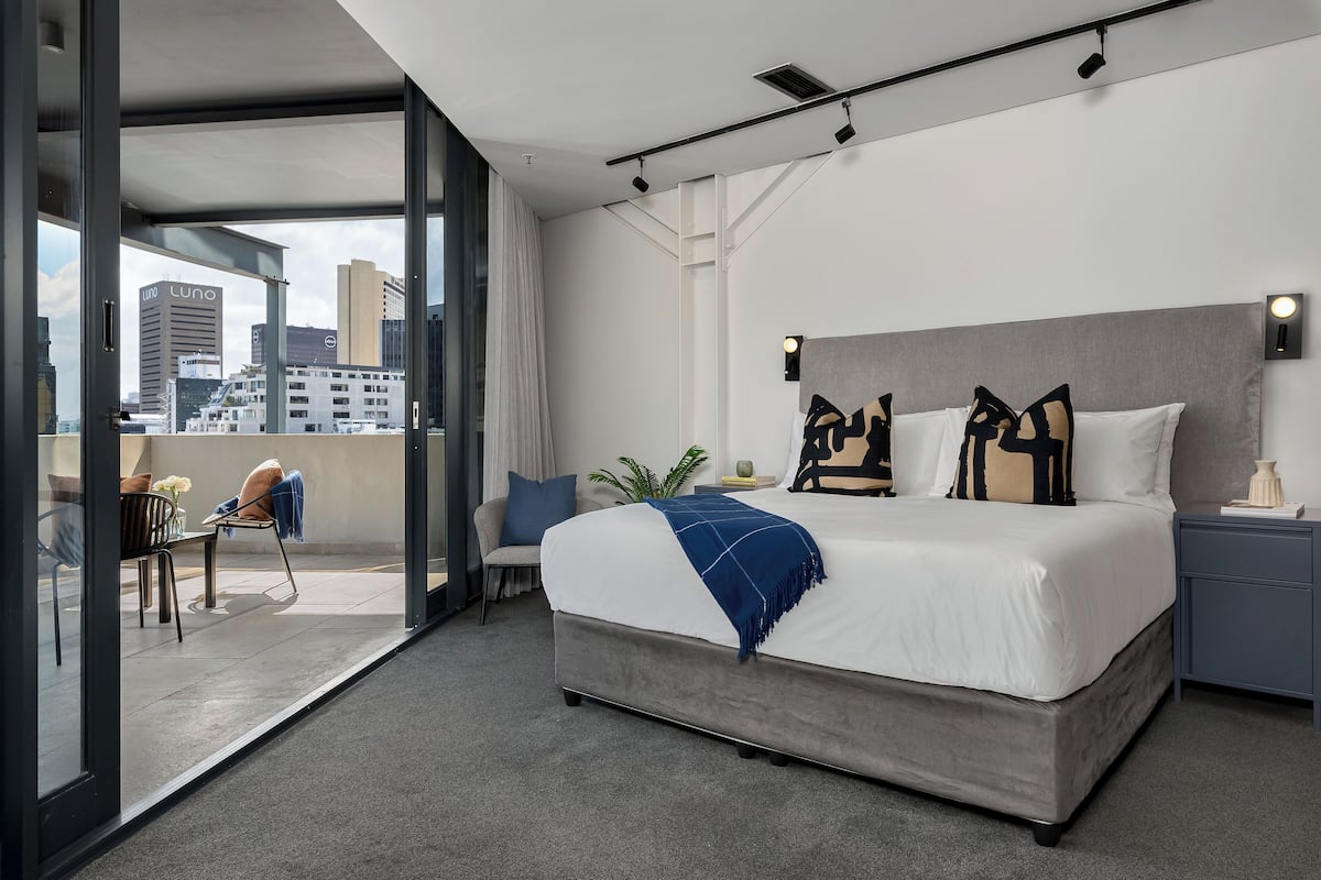 Quarto de hotel moderno com uma cama grande, uma escrivaninha e vista para a cidade através de janelas do chão ao teto.