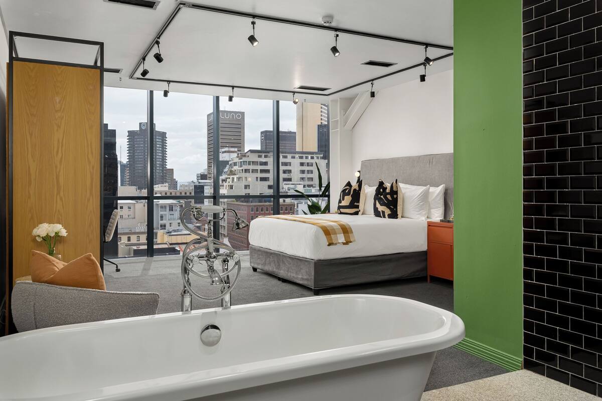 Quarto de hotel moderno com uma cama grande, uma banheira e vista para a cidade através de amplas janelas.
