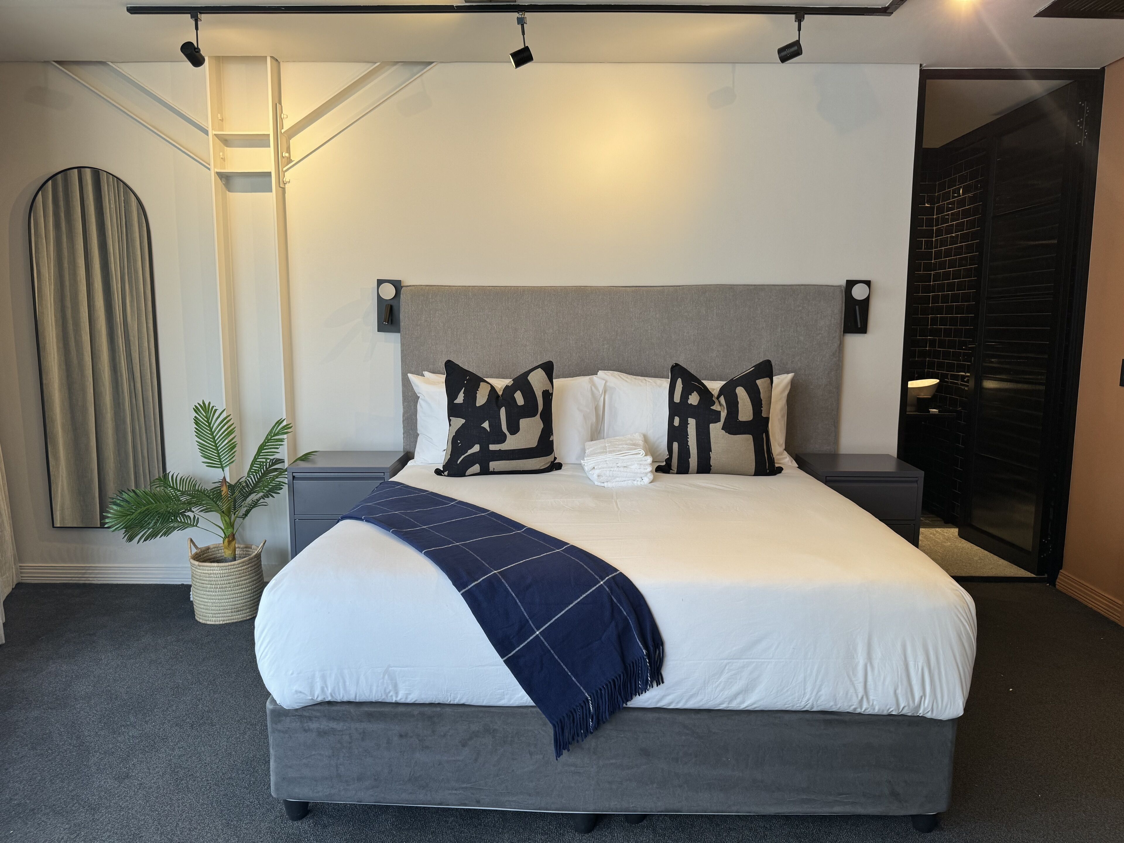 A modern hotel room with a large bed, grey headboard, and a potted plant.