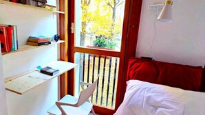 Ein Zimmer mit einem Bett, einem Schreibtisch mit Büchern, einem Stuhl, einem Fenster mit Blick auf Bäume und einer roten Wand.