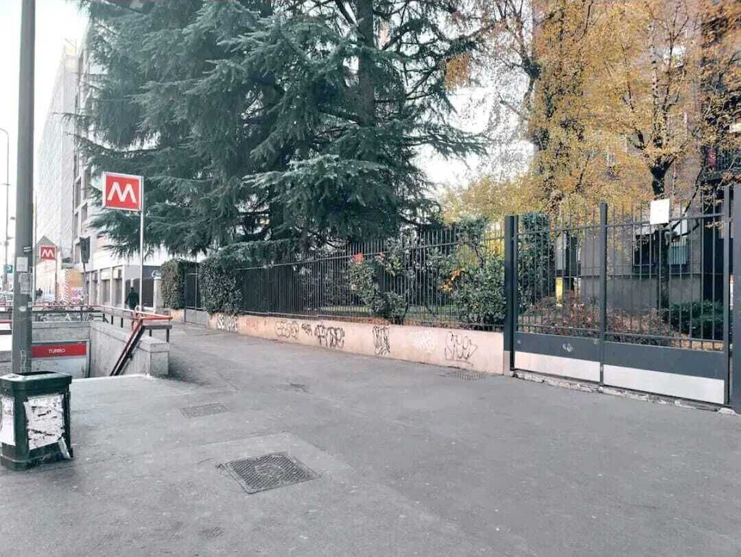 A paved area with a metal fence, trees, and a building with a red 'M' sign.