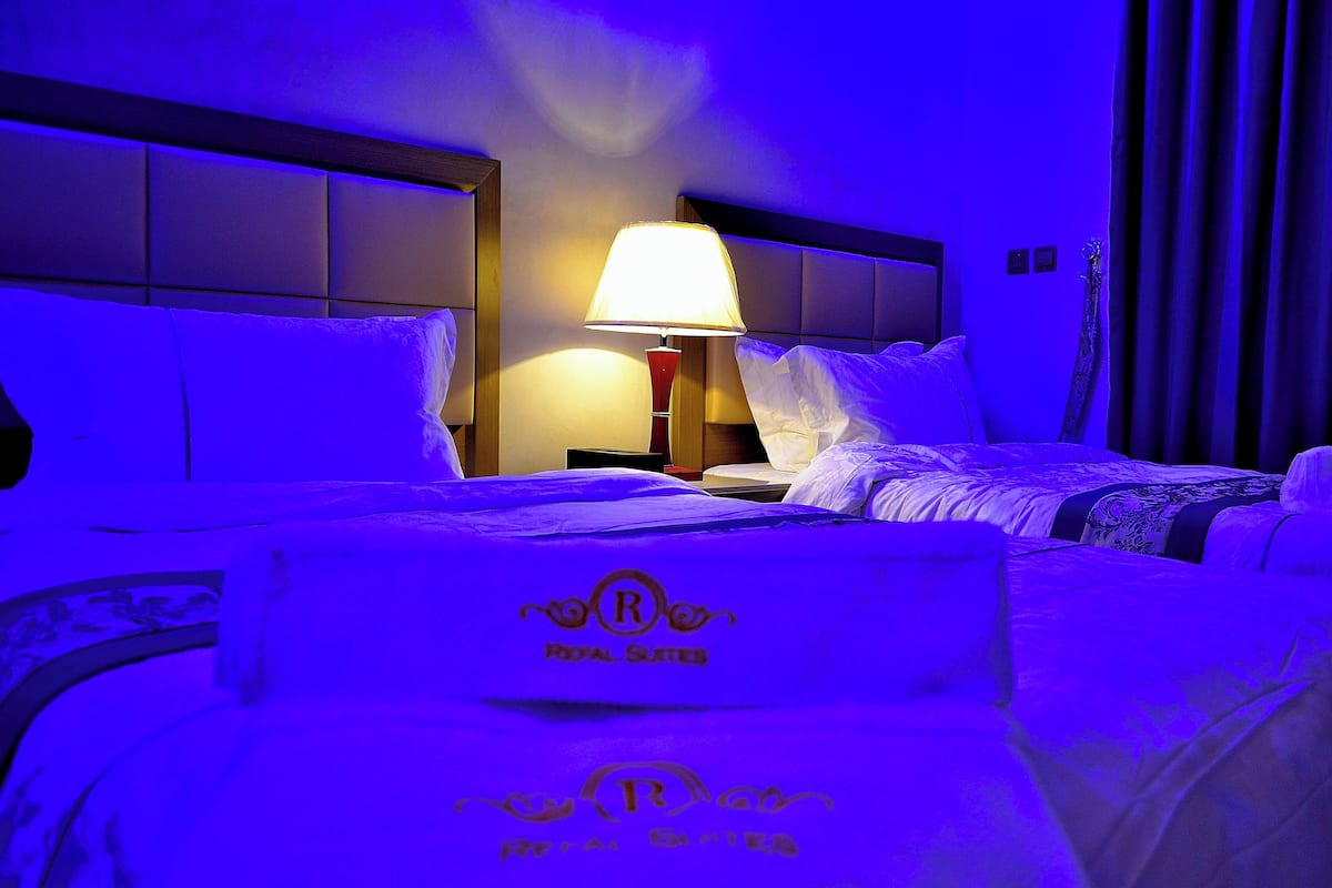 Une chambre d’hôtel avec deux lits, une table de chevet avec une lampe et une tête de lit ornée d’un logo.