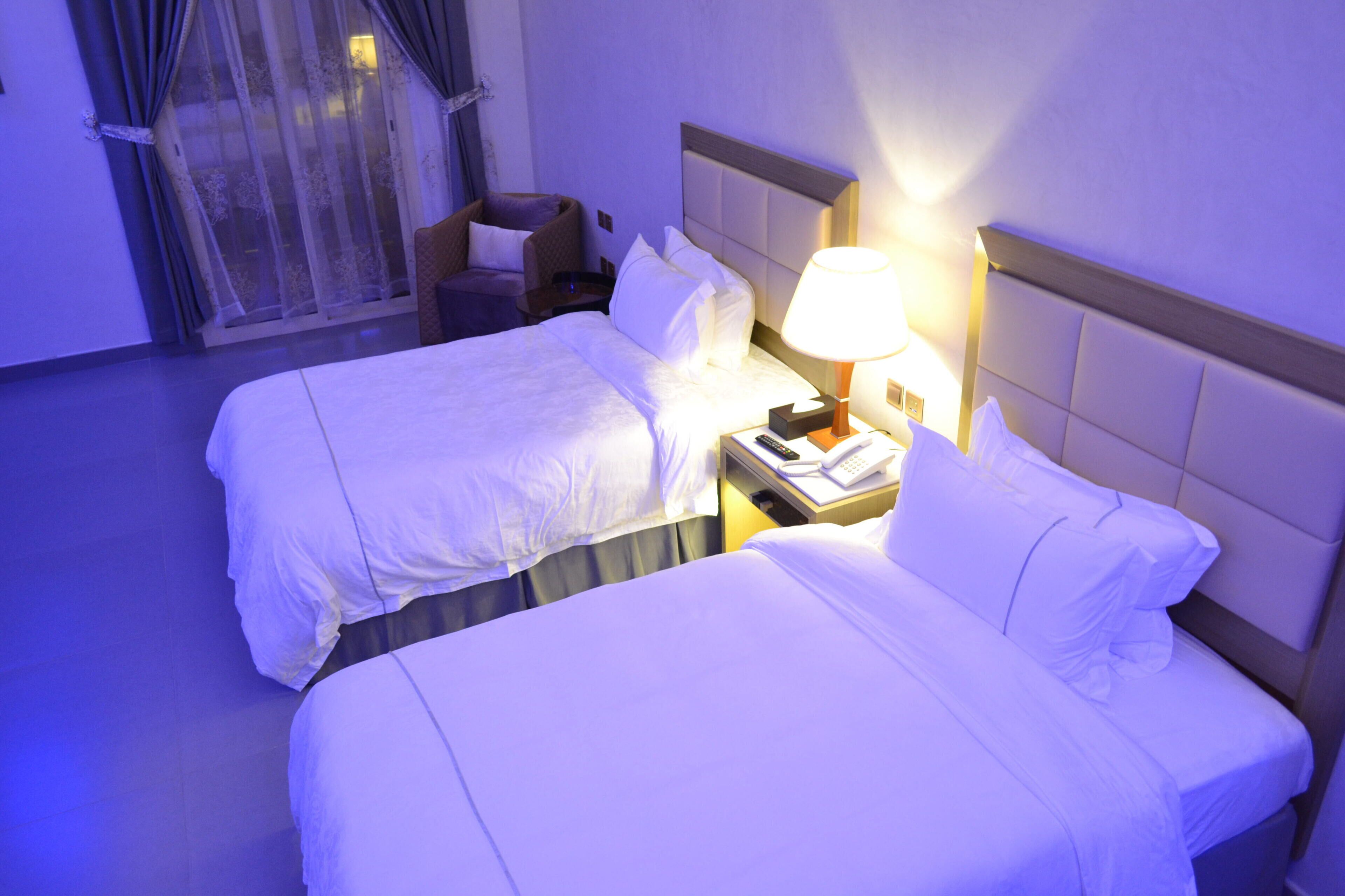 Une chambre d’hôtel avec deux lits, une table de chevet avec une lampe, une chaise et une fenêtre avec des rideaux.