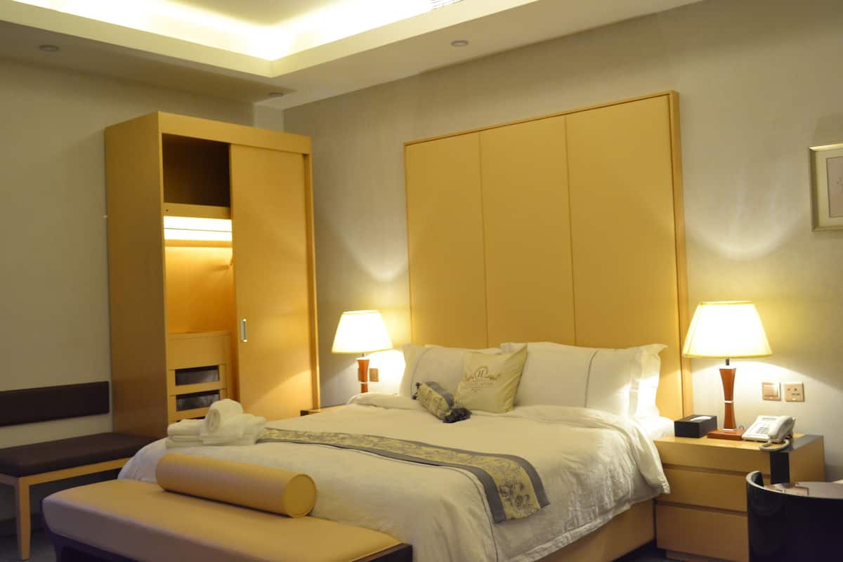 Une chambre d’hôtel avec un grand lit, deux lampes de chevet, un téléphone sur la table de chevet, une table de chevet, un banc et une armoire.