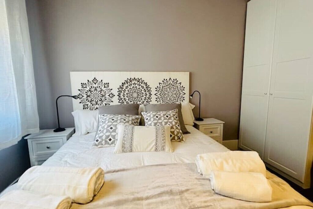A neatly made bed with white linens and multiple pillows, a bedside table with a lamp, and a wardrobe in a hotel room.
