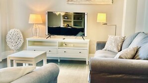 A living room with a flat-screen TV on a white TV stand, a grey sofa, a small white side table, and two floor lamps. - Aparta 4 Habitaciones Terrassa Centro, Parking (Terrassa)