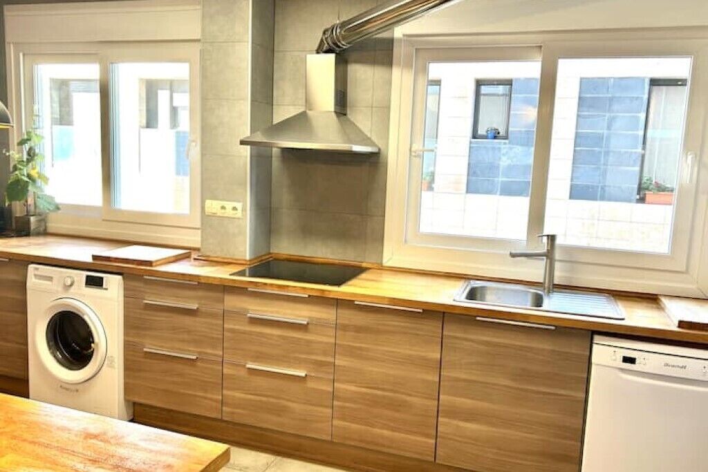 A modern kitchen with wooden cabinets, a stainless steel range hood, a built-in dishwasher, and a large window providing natural light.