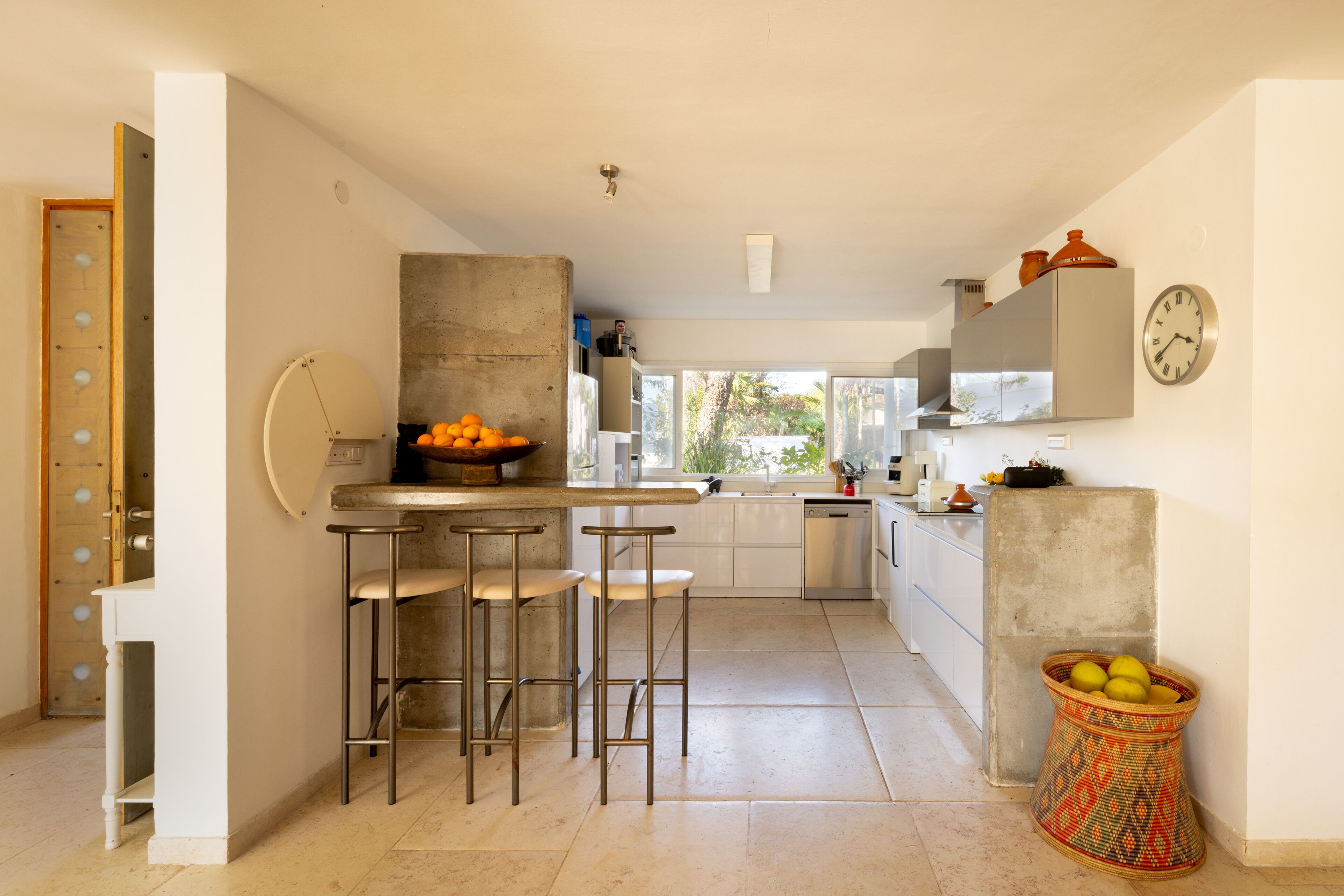 Une cuisine moderne avec un îlot central en béton, des tabourets de bar, une grande fenêtre et une horloge au mur.