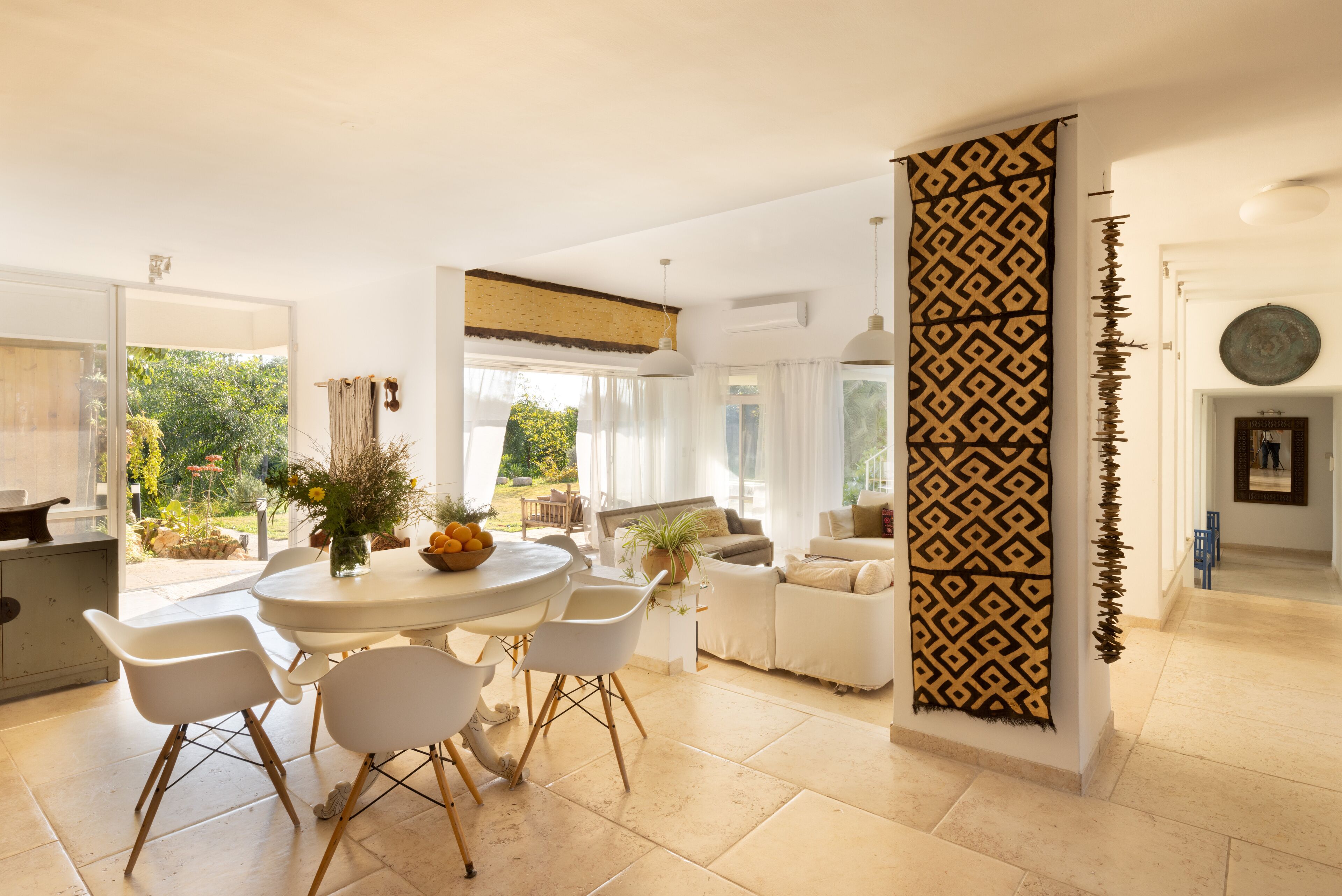 A bright, open-plan living area with a dining table, chairs, and a sofa. A patterned wall divider is present, and there is a view of greenery outside.