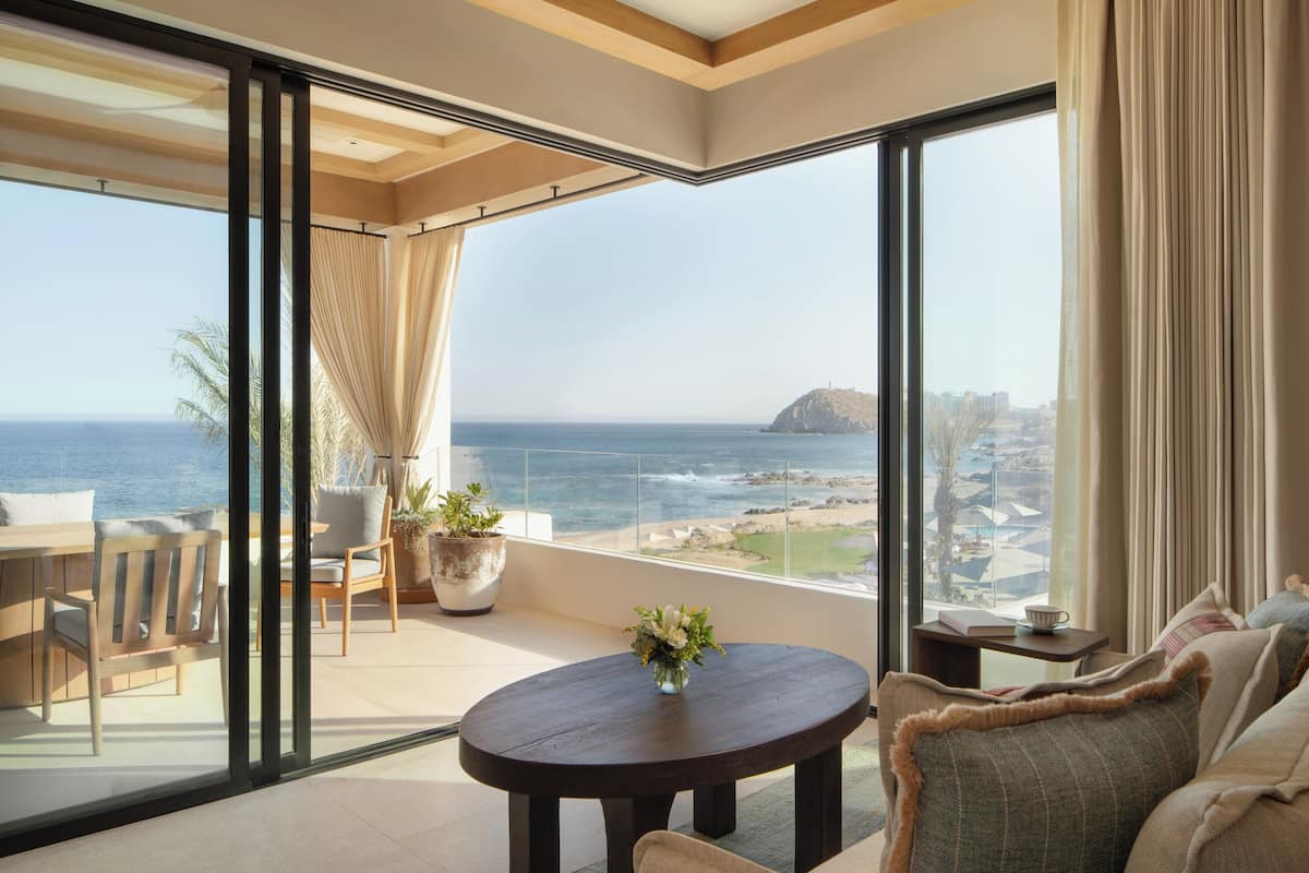 A balcony with a view of the ocean and a distant island. There is a round table with a vase of flowers and a sofa with cushions.