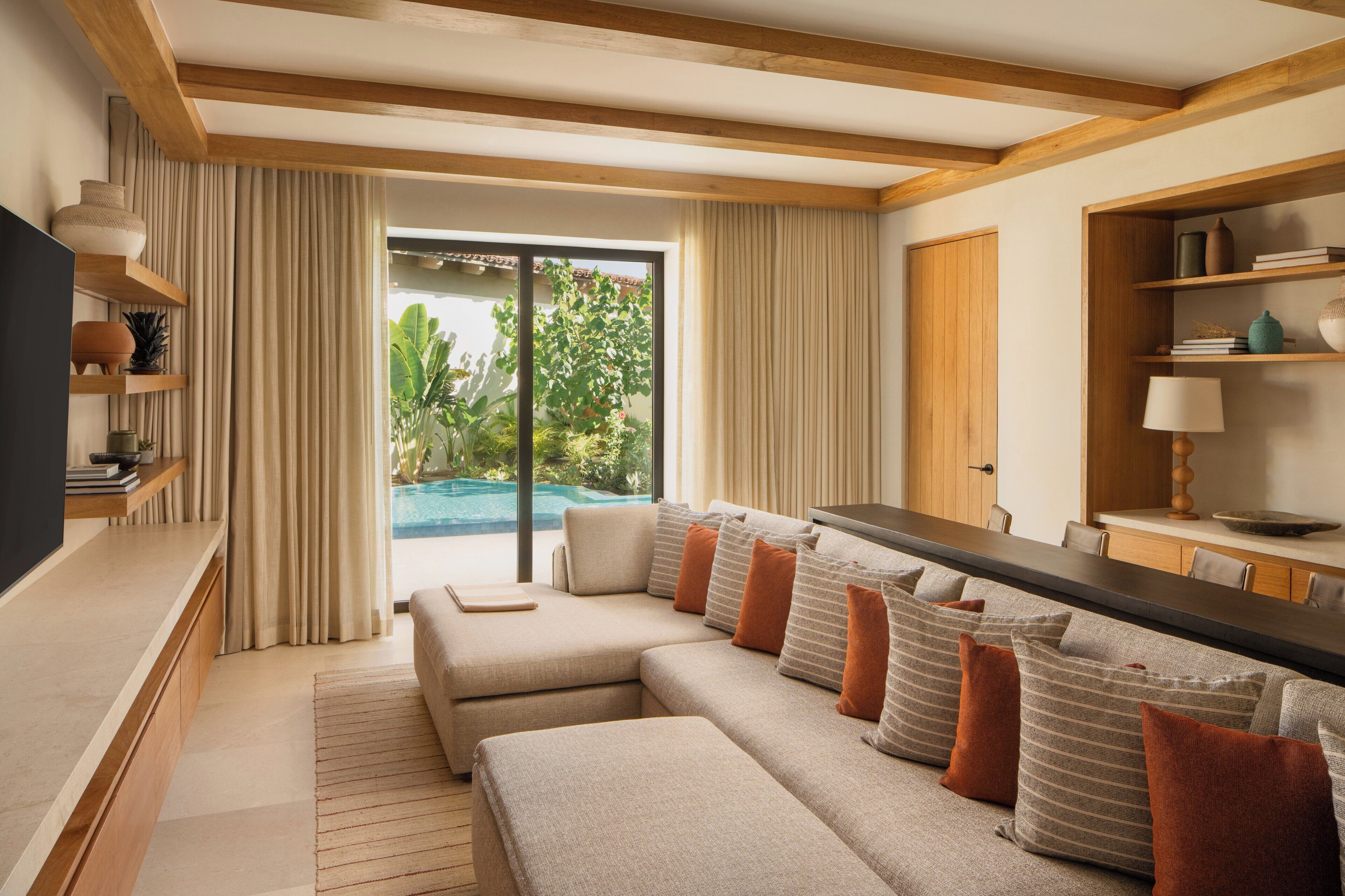 A modern living room with a sectional sofa, wooden beams, a TV, and a view of a swimming pool.