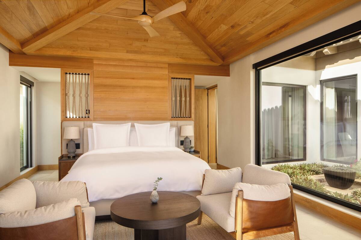 A modern hotel room with a large bed, two bedside tables, a round coffee table, and a seating area with two chairs.