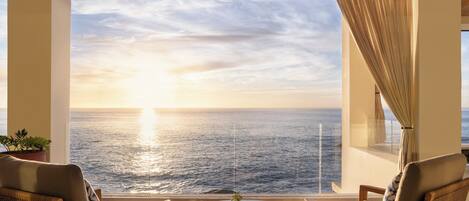 A room with a view of the ocean at sunset, featuring a round table and two chairs.