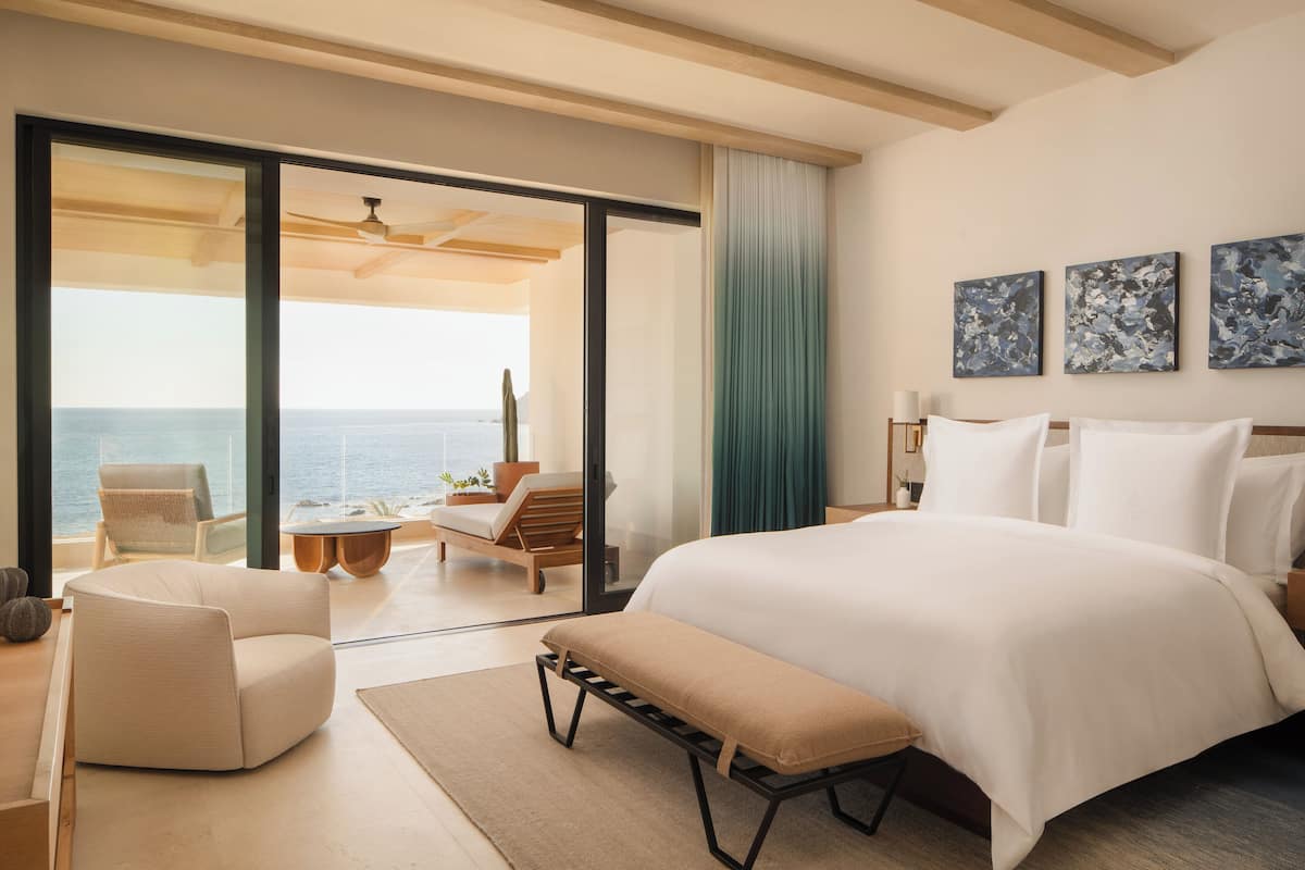 A modern hotel room with a large bed, a seating area with a view of the sea, and artwork on the wall.