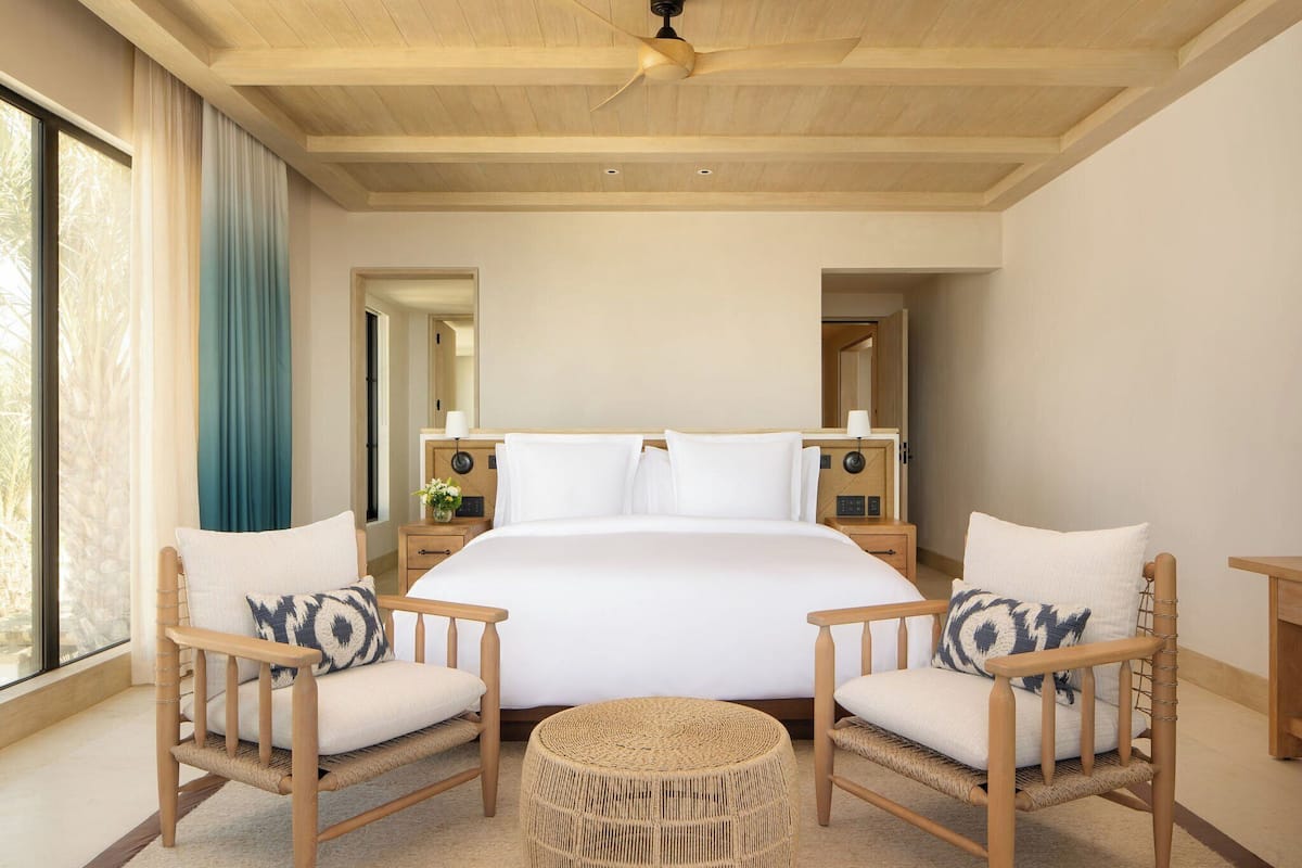 A spacious bedroom with a large bed, two wooden chairs with cushions, a round woven ottoman, and a ceiling fan.
