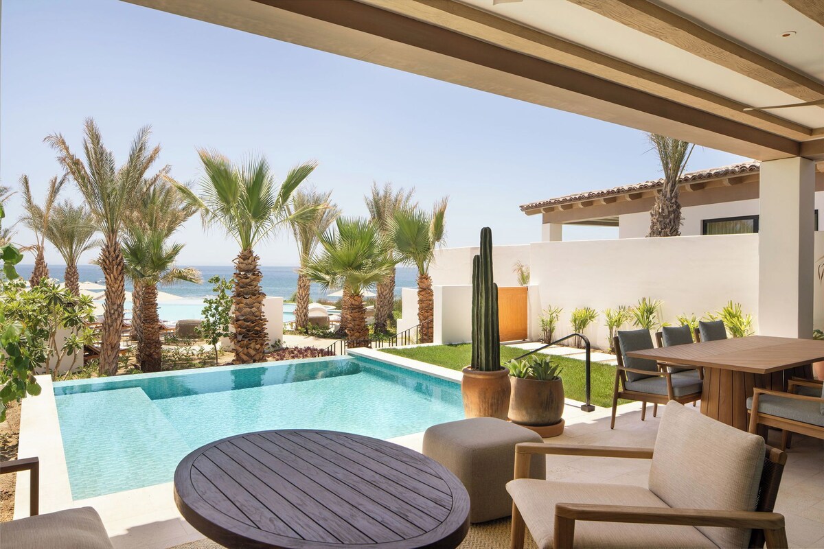 An outdoor pool area with a view of the ocean, surrounded by palm trees and a wooden pergola.