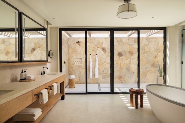 Un baño moderno con una bañera grande, un tocador de madera, una pared de piedra y un divisor de vidrio corredera que da a la zona de ducha.